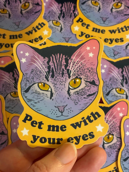 Sticker of a cat with yellow eyes and text 'Pet me with your eyes' held by a hand.