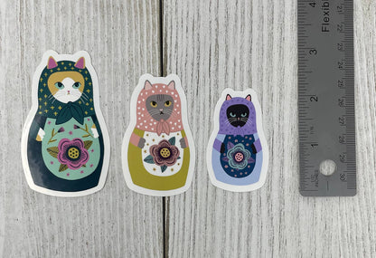 Three colorful cat-themed stickers on a wooden surface with a ruler for scale.