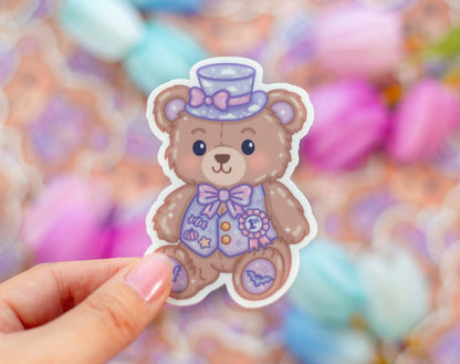 Sticker of a teddy bear with a top hat and bow tie held by a hand against a colorful blurred background.